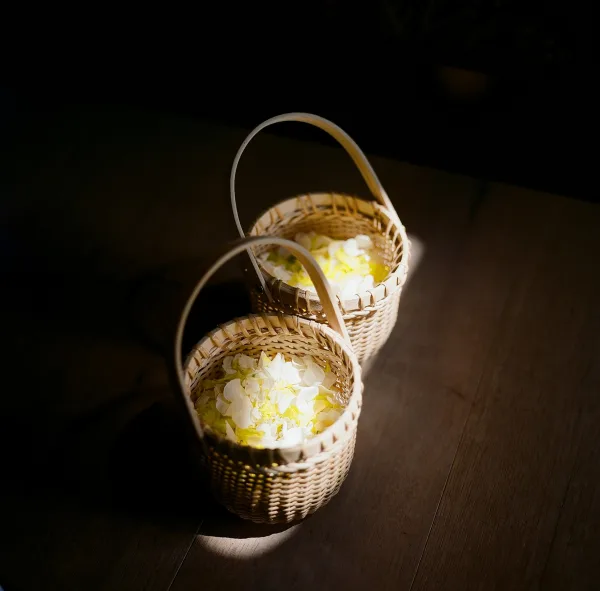 Flower girl baskets with white and yellow flower petals on a sunlit wood floor, woven wicker details casting soft shadows