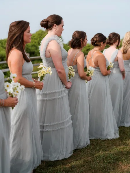 Bridesmaids lineup in gray bridesmaid dresses holding small white daisy bouquets on an outdoor lawn by a wooden fence, low bun updos