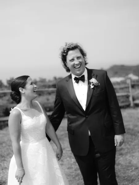 Couple portrait in a black and white wedding photo, bride in strapless dress and groom in tuxedo laughing by a rustic fence field
