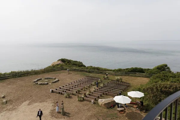 Ceremony setup with outdoor ceremony seating on wood benches, aisle florals and wine barrels facing a stone circle altar on an ocean-view bluff