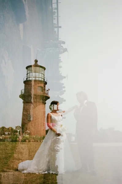 Couple portrait of bride and groom holding hands, her veil and bouquet flowing beside a lighthouse in a grassy coastal field