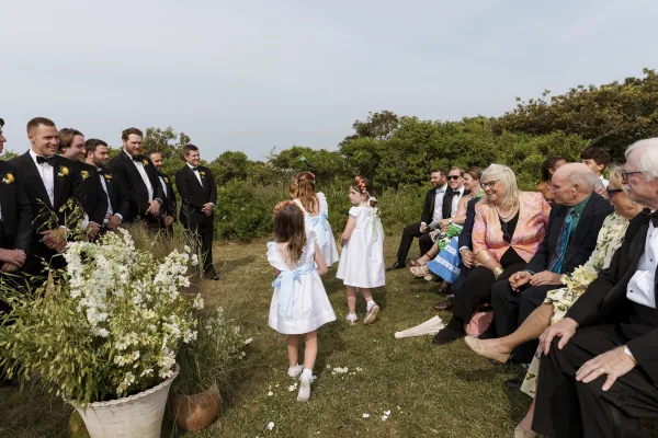 Wedding processional with flower girl walking down aisle in a white dress with blue sash and floral crown on a sunny outdoor lawn aisle