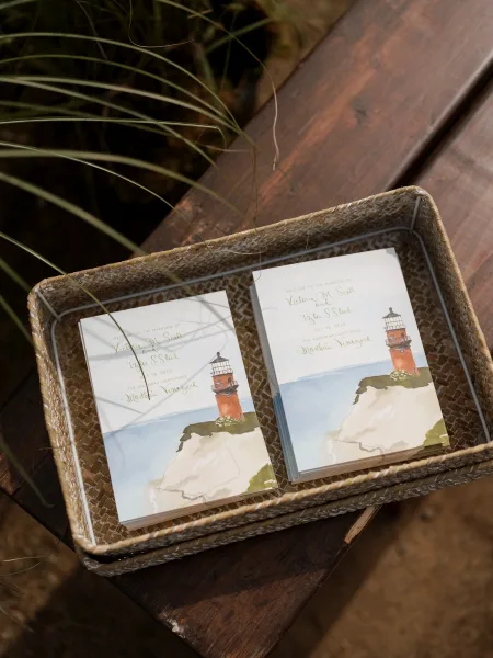 Wedding programs with ceremony program design featuring a lighthouse illustration, stacked in a woven basket on a wooden bench outdoors
