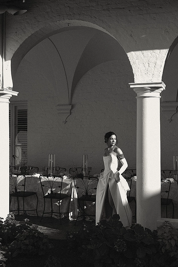 Bridal portrait of a bride in long gloves wearing a strapless slit gown beside a banquet table under an arched courtyard colonnade