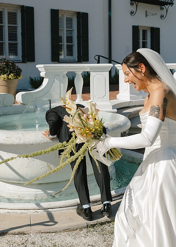 Wedding couple moment as the groom splashes in a fountain while the bride laughs, holding a bouquet beside a white balustrade courtyard