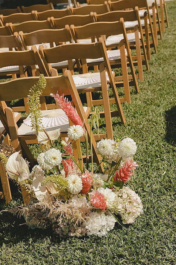 Ceremony aisle decor with outdoor ceremony seating, wood folding chairs lined with paper fans and lush floral ground arrangements on sunlit grass lawn