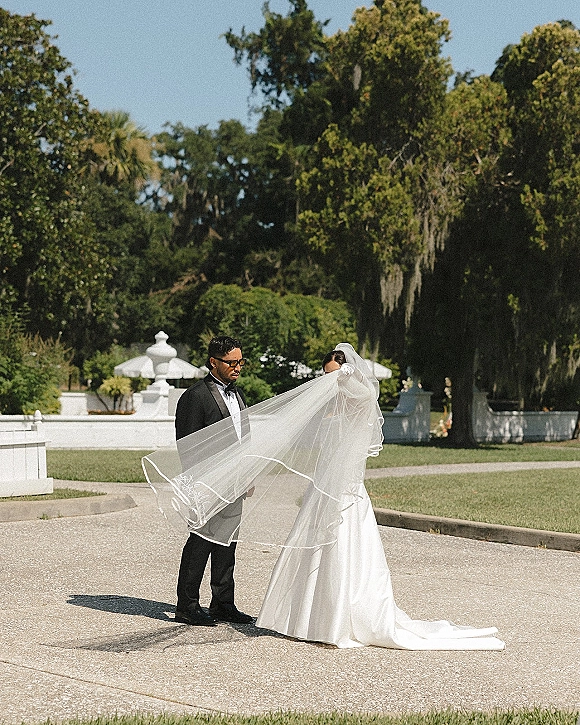 Couple portrait of bride and groom portrait with the bride’s long veil draped over him, in tuxedo and sunglasses on a sunny garden lawn