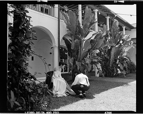 Wedding candid moment as groom adjusting bride shoe while she sits in strapless gown, suspenders and string lights in stucco courtyard