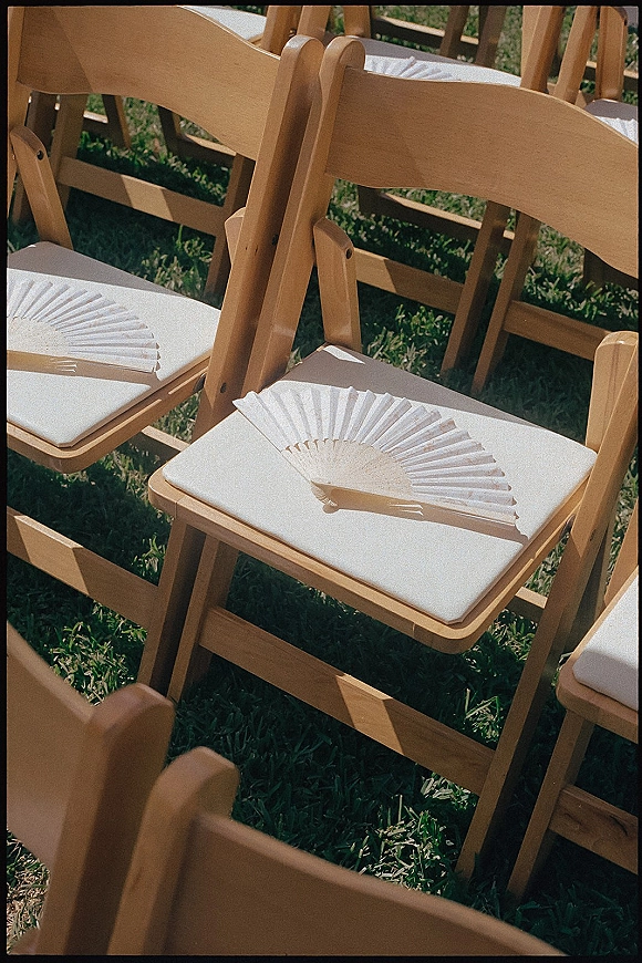 Ceremony seating with outdoor ceremony chairs, wood folding chairs with white cushions and paper hand fans on a sunlit lawn