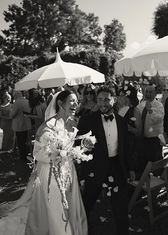 Wedding recessional as bride and groom walk up the aisle laughing, bouquet and veil flowing while guests toss flower petals in a garden setting