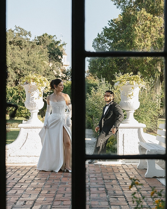 Couple portrait of bride in strapless dress and opera gloves with groom in tux and sunglasses on a brick patio by stone balustrade