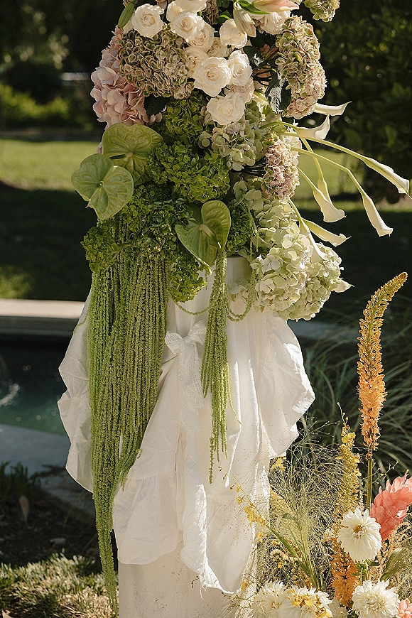 Ceremony floral arrangement of white roses, hydrangea, and hanging amaranthus on a fabric-draped pedestal beside meadow blooms on a garden lawn