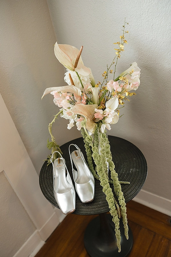 Bridal shoes and silver wedding heels beside a sculptural bouquet of calla lilies, anthurium, and orchids on a black pedestal table