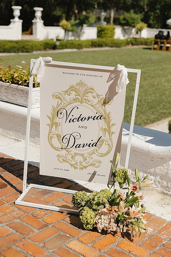 Wedding welcome sign on a white stand with lace ribbon accent and hydrangea, lilies, and tulips on a brick patio by hedges
