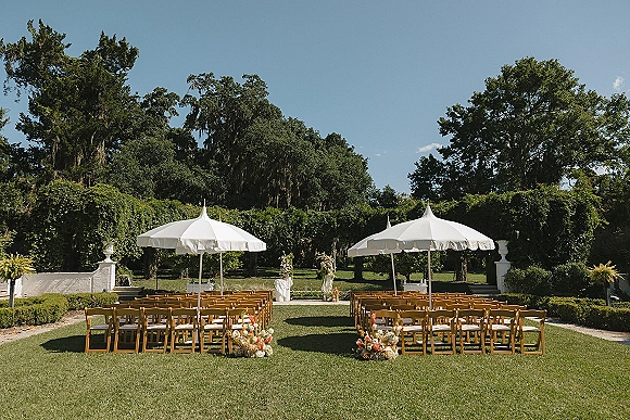 Ceremony setup for an outdoor wedding ceremony with wood chairs, white patio umbrellas, and aisle flowers on a sunny garden lawn