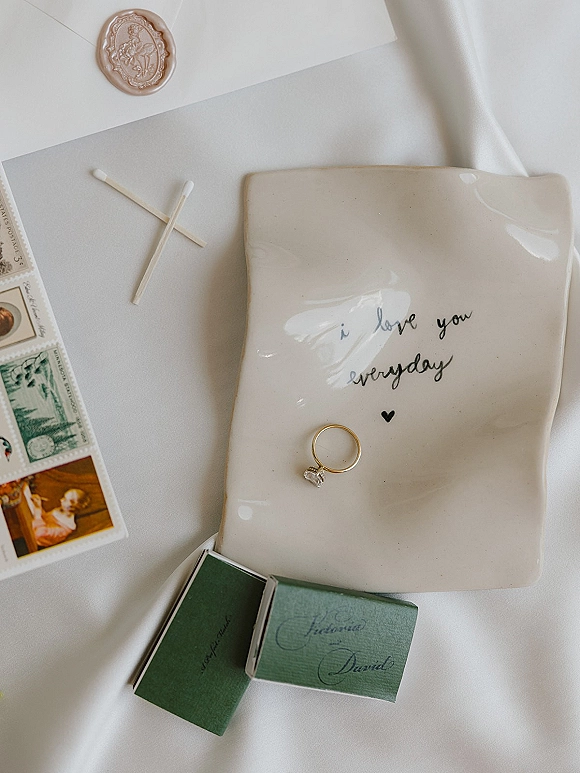 Wedding rings and engagement ring flat lay on a ceramic ring dish with diamond solitaire, calligraphy, wax seal, and stamps on white fabric