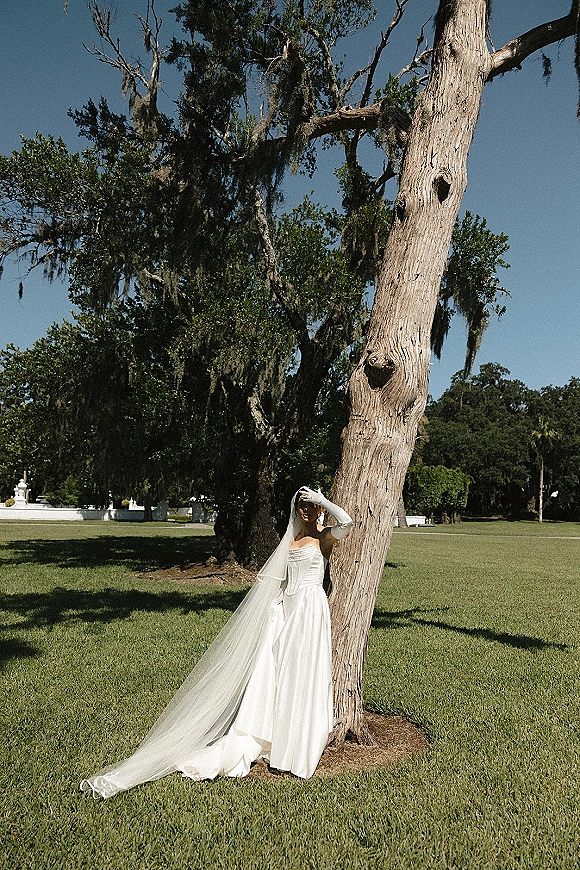 Bridal portrait of a bride leaning on a large tree, wearing a strapless satin gown, long veil train, and opera gloves in a sunny park lawn