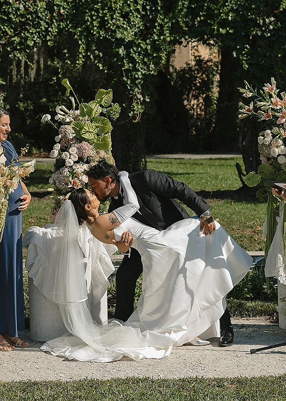 Wedding kiss as groom dips the bride in a strapless gown, veil flowing, beside floral pillars on a garden aisle