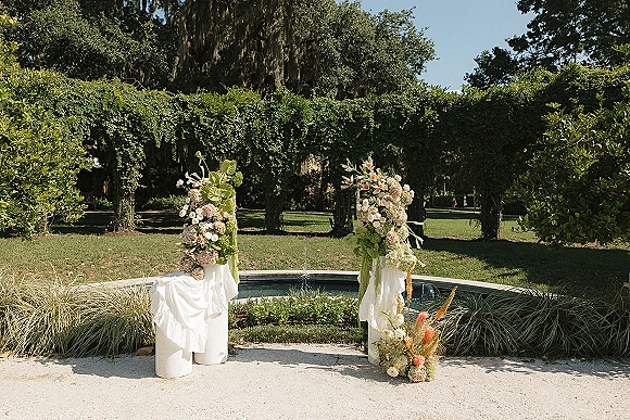 Ceremony floral pillars with wedding floral pillars of pastel blooms on pedestal stands, white draped fabric and ribbon, set on a garden lawn by a fountain