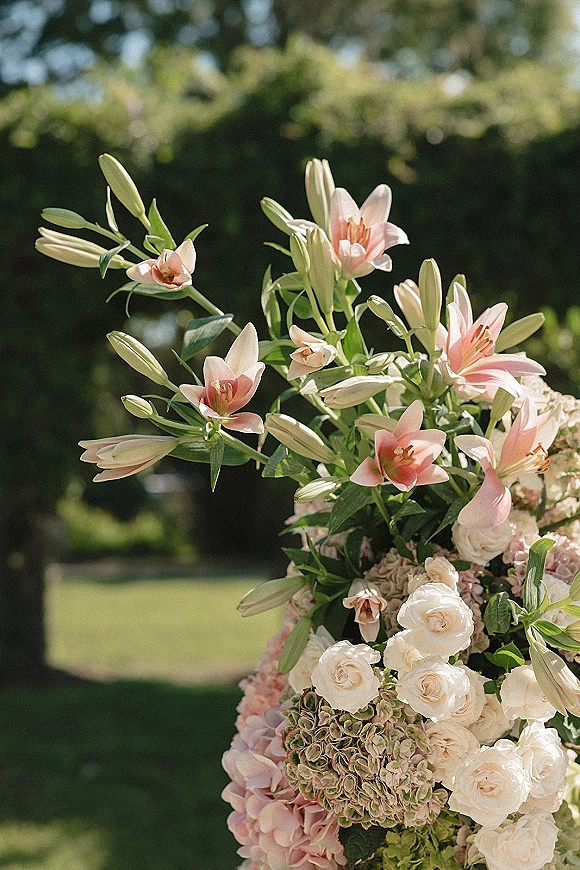 Wedding florals with pink lily wedding flowers, white roses and hydrangea with greenery, arranged outdoors on a sunlit garden lawn with trees