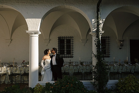 Wedding kiss portrait of bride and groom kissing in a white-column courtyard, bride in strapless gown and opera gloves, ivy accents nearby