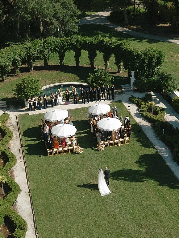 Outdoor wedding ceremony with white parasol shade as the bride walks the aisle past chairs and florals, poolside on a manicured lawn