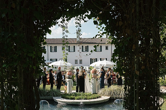 Outdoor wedding ceremony with bride and groom at the altar, officiant and guests on an estate lawn by a fountain under blue sky