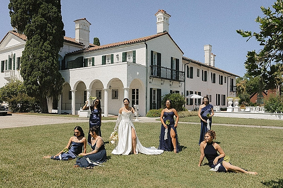 Bridal party portrait of bride with bridesmaids in navy dresses holding white bouquets on a villa lawn under arched colonnade