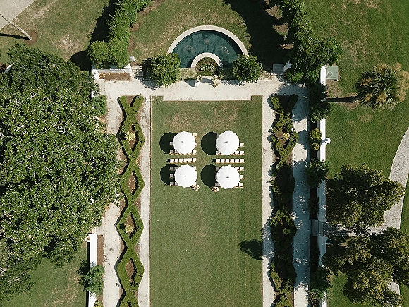Outdoor ceremony setup with an aerial wedding ceremony view of wooden chair rows, aisle flowers, and white patio umbrellas on a manicured lawn near a fountain