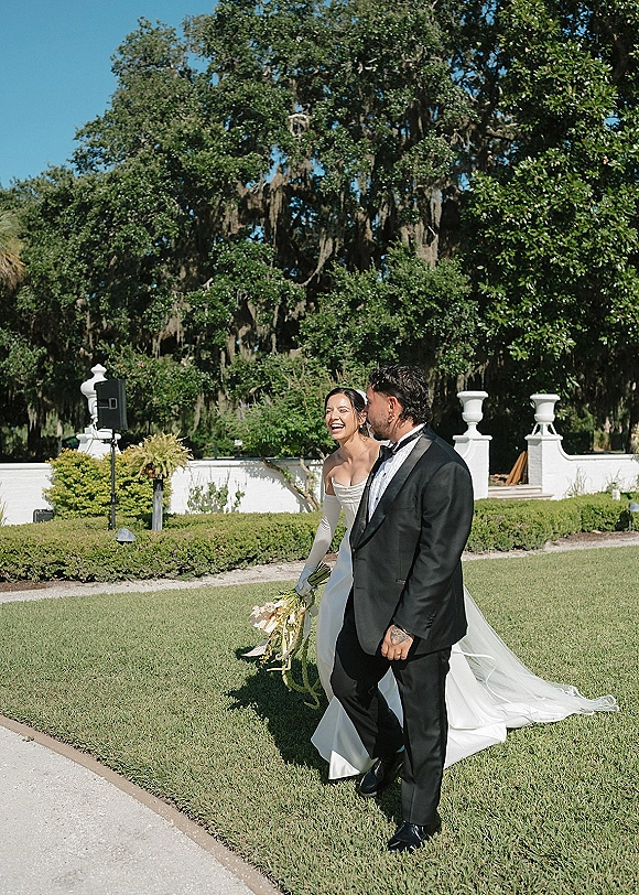 Couple portrait of bride and groom walking, bride in strapless dress and long gloves holding bouquet on garden lawn by white wall