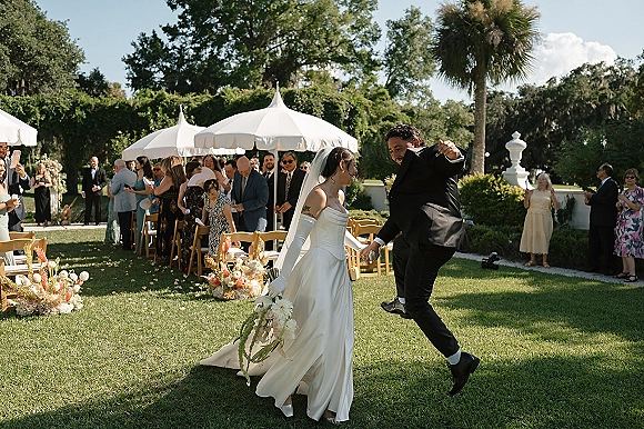 Wedding recessional as bride and groom exit hand in hand, bouquet and veil flowing, guests cheering along a sunny garden aisle with umbrellas