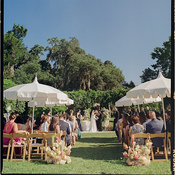 Outdoor wedding ceremony with bride and groom under a floral arch, guests in wooden chairs on a garden lawn under white umbrellas.