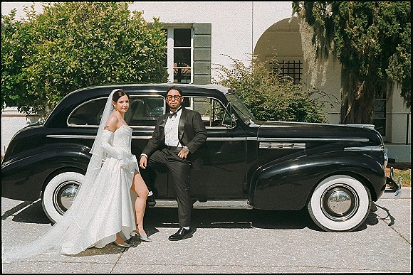 Couple portrait of bride and groom with car, leaning on a black vintage getaway car in sunglasses outside a stucco venue driveway