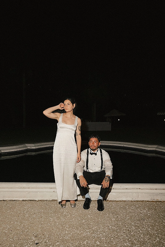 Couple portrait at a nighttime wedding portrait by the pool, bride standing in a simple dress and groom sitting in suspenders under the night sky