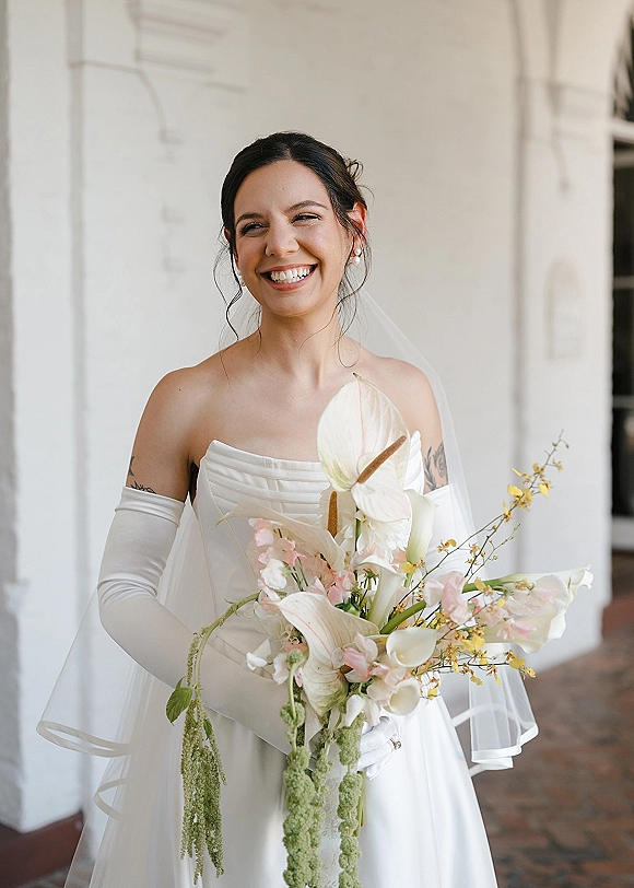 Bridal portrait of a smiling bride in a strapless wedding dress with veil and long gloves, holding a bouquet by an arched doorway