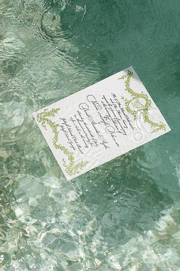 Wedding invitation with calligraphy wedding invitation text and green botanical border floating on rippling water with sunlit reflections