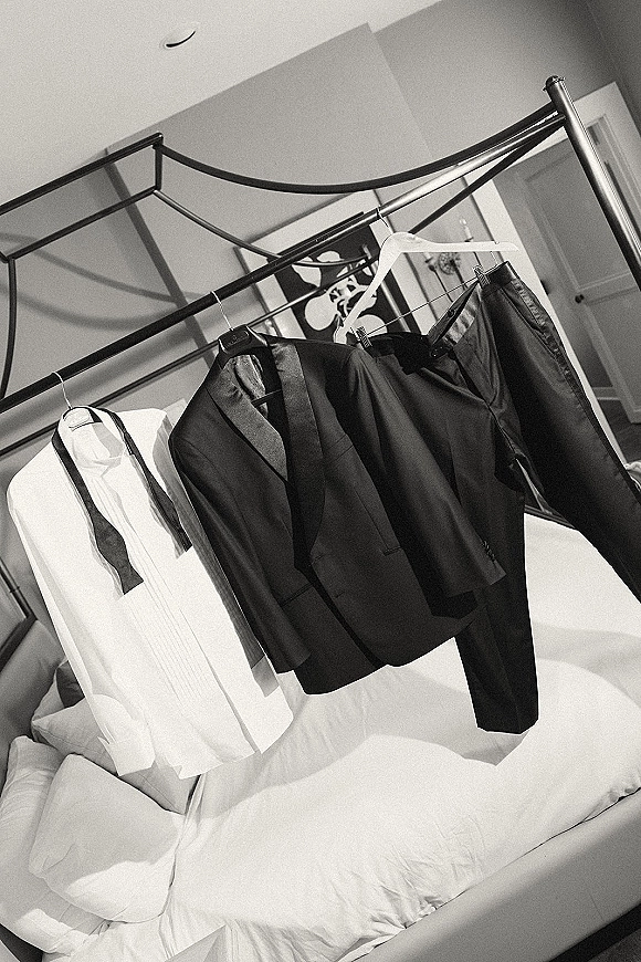Groom attire, wedding tuxedo details displayed on hangers above a bed, with tuxedo jacket, bow tie, shirt, and trousers in a bedroom