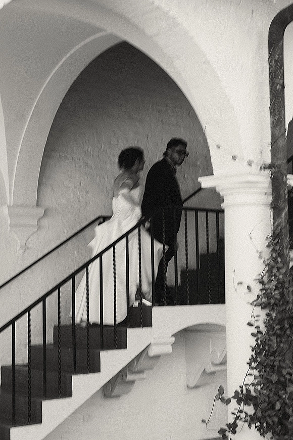 Wedding couple portrait in black and white, bride in veil and strapless dress following groom in glasses up an arched staircase walkway