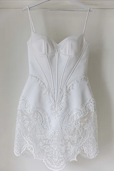 Wedding dress with lace applique corset bodice, beading, and spaghetti straps hanging on a hanger against a neutral wall