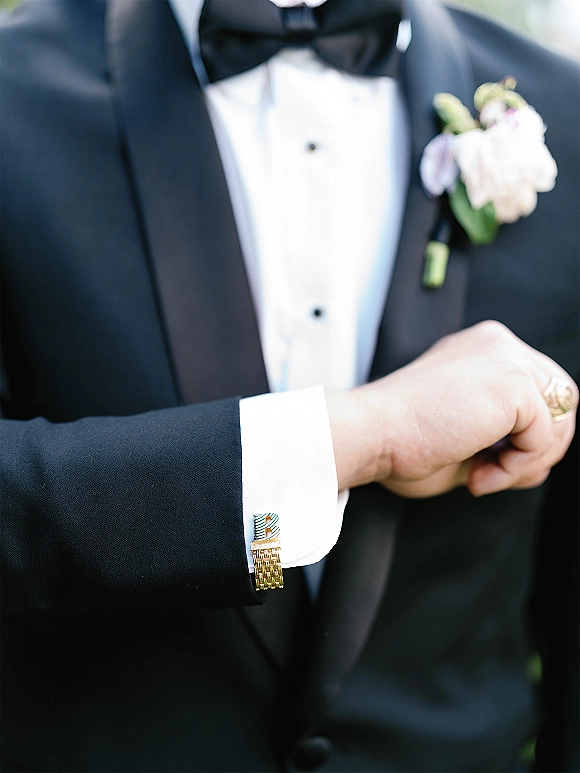 Groom tuxedo details with wedding cufflinks, black bow tie and boutonniere on white shirt, soft outdoor greenery blurred behind