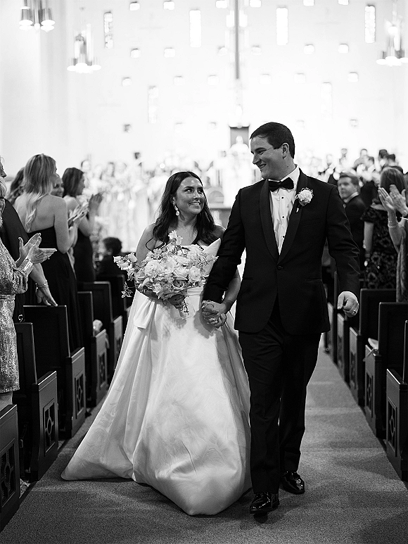 Wedding recessional as bride and groom hold hands walking the church aisle, bride with bouquet and gown train, guests clapping beneath chandeliers