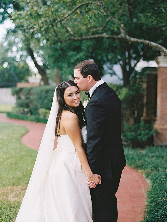 Couple portrait of groom kissing bride’s forehead as they hold hands, her veil flowing in a garden by a brick walkway and trees