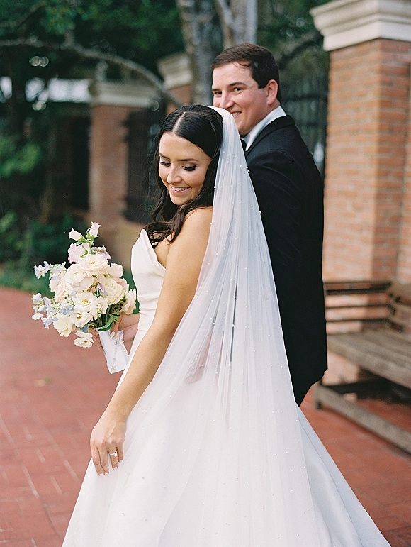 Couple portrait of bride holding bouquet, groom behind her, with a long wedding veil along a brick walkway under an archway