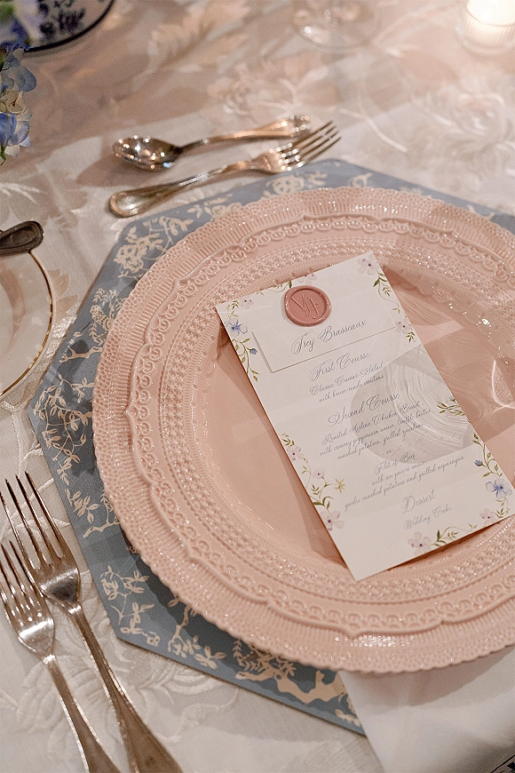 Wedding tablescape with blush wedding tablescape details, beaded charger, pink plate, wax-seal menu, blue placemat, candle, and florals on damask cloth