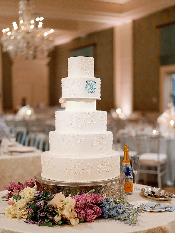 Wedding cake on a silver cake stand, five-tier white design with embossed icing and monogram topper beside champagne in warm reception room