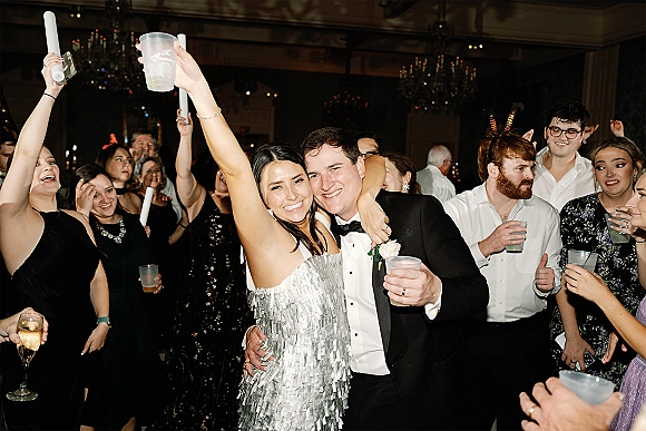 Wedding reception dance as groom in tuxedo and bride in strapless silver sequin dress raise drinks amid glow sticks under chandeliers