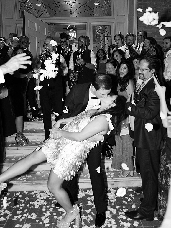 Wedding exit kiss as groom dips bride in a short fringe dress on steps, confetti petals falling amid cheering guests by lantern-lit doorway