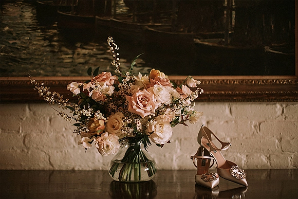 Wedding details flat lay with bridal shoes close up, jeweled buckle heels beside roses and baby's breath in a green glass vase on wood tabletop
