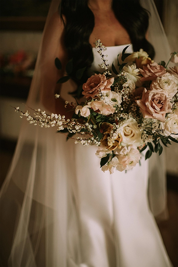 Bridal bouquet of blush and ivory bouquet roses with white filler flowers and greenery, held against a strapless dress in soft indoor light