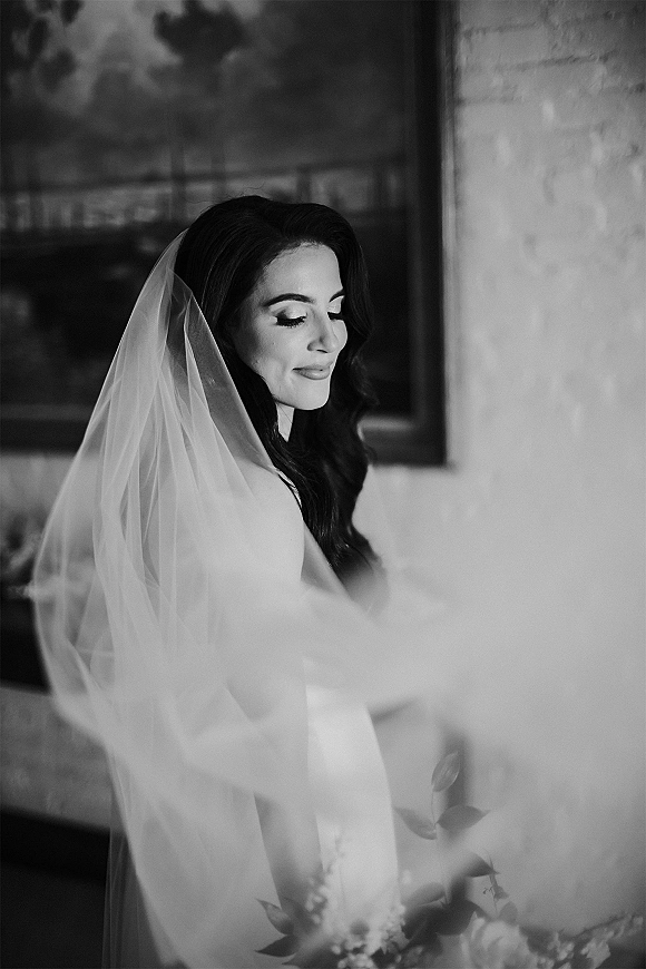 Bridal portrait in black and white of a bride in veil looking down, long veil over her shoulders, posed indoors by a brick wall with framed artwork